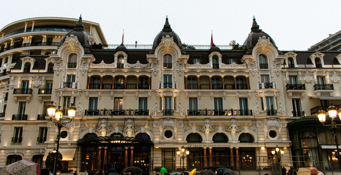 The grand, ornate Beaux-Arts facade of the Hotel de Paris in Monaco on a rainy day, with people carrying umbrellas in the foreground.