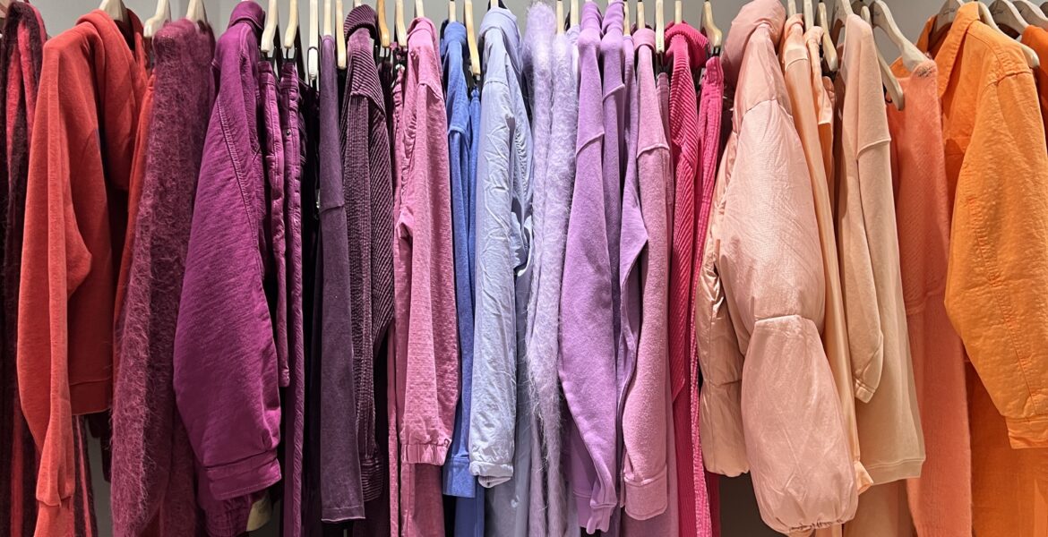 A vibrant clothing rack displaying key summer 2026 colours organized in a gradient from red and purple to peach and orange.