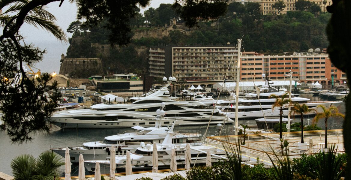 Luxury mega yachts docked in Monaco's Port Hercules, framed by Mediterranean greenery and iconic Monte Carlo architecture during the Monaco GP Fashion 2026 showcase.