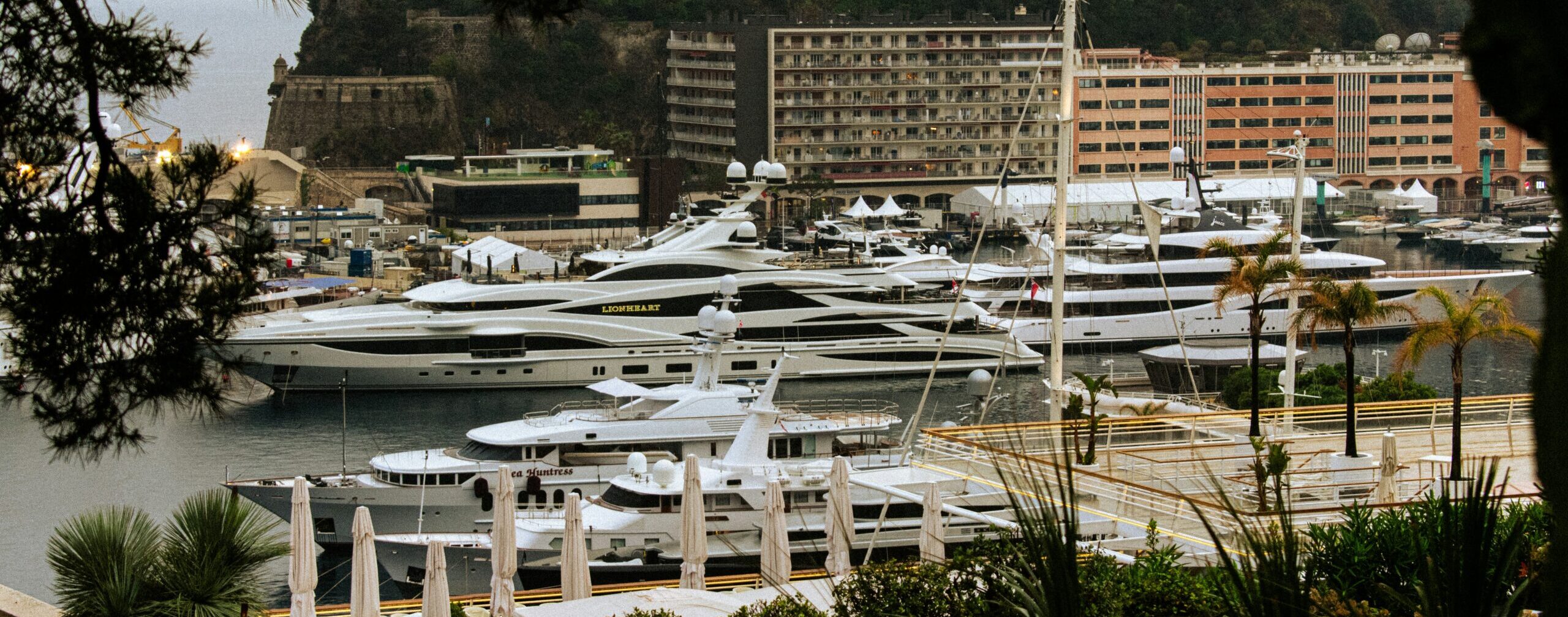 Luxury mega yachts docked in Monaco's Port Hercules, framed by Mediterranean greenery and iconic Monte Carlo architecture during the Monaco GP Fashion 2026 showcase.