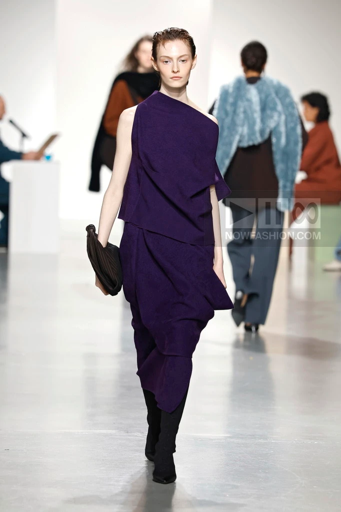 Model on the runway at the CFCL fashion show in Paris, Fall Winter 2026 Ready To Wear Fashion Week, Runway Look #010 - CFCL Fall/Winter 2026 runway look featuring an asymmetrical deep purple draped knit dress.