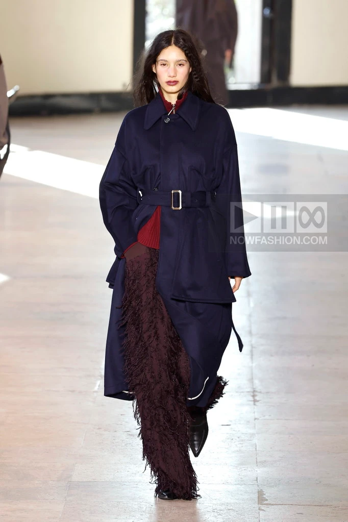 Model on the runway at the Ujoh fashion show in Paris, Fall Winter 2026 Ready To Wear Fashion Week, Runway Look #009 - Ujoh Fall/Winter 2026 runway look combining a navy belted coat, burgundy top, and dark purple fringed trousers.
